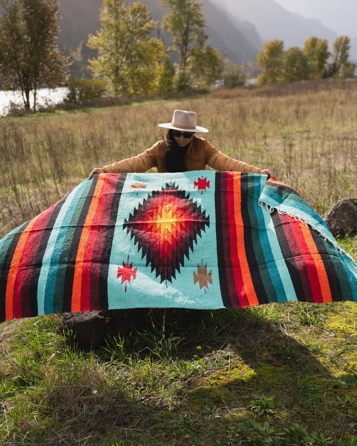 Woman wearing a cowboy hat holding a teal and red handwoven mexican diamond blanket in front of sunset in the mountains
