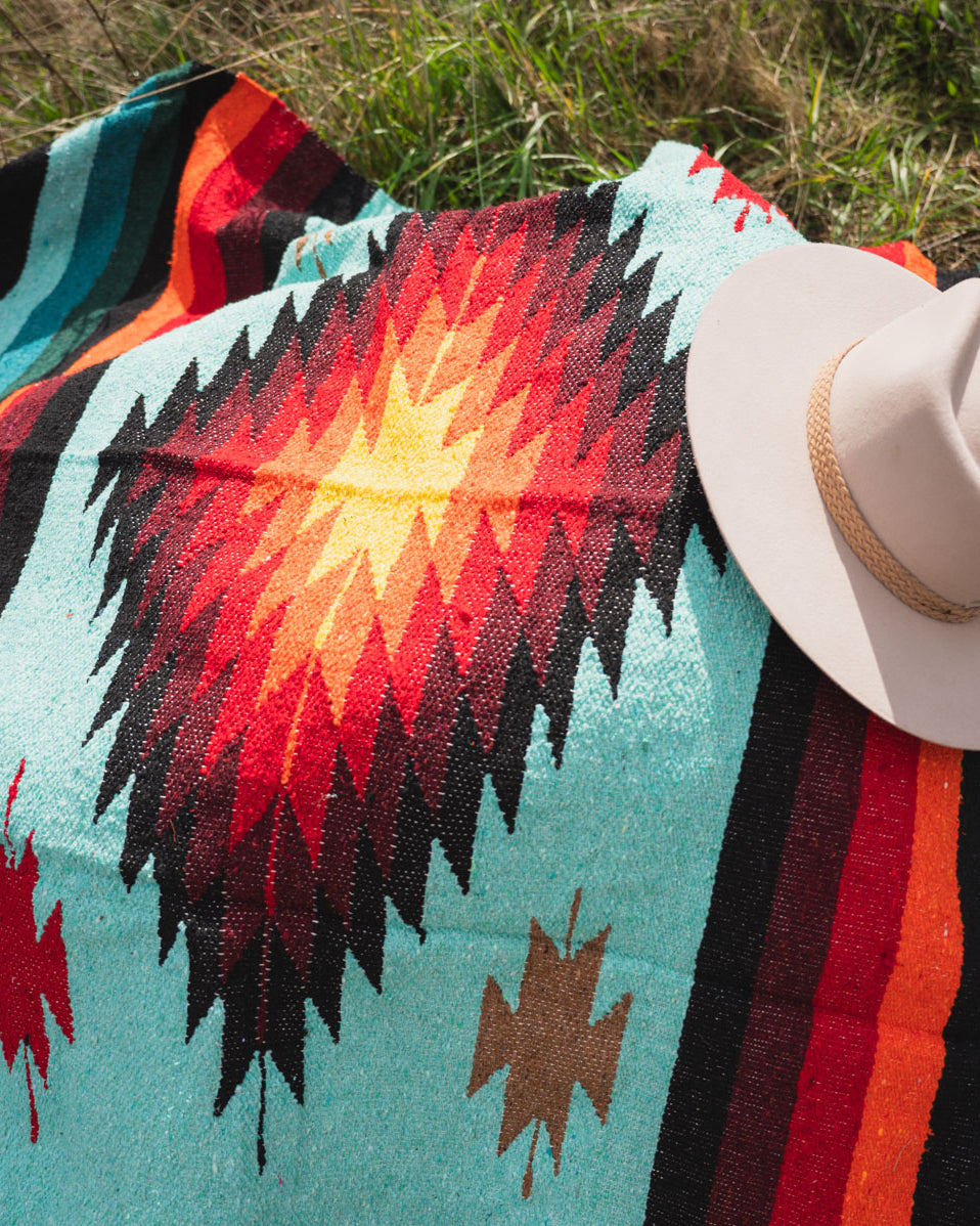 teal and red handwoven diamond mexican blanket laying on the ground with a cowboy hat on top of it.