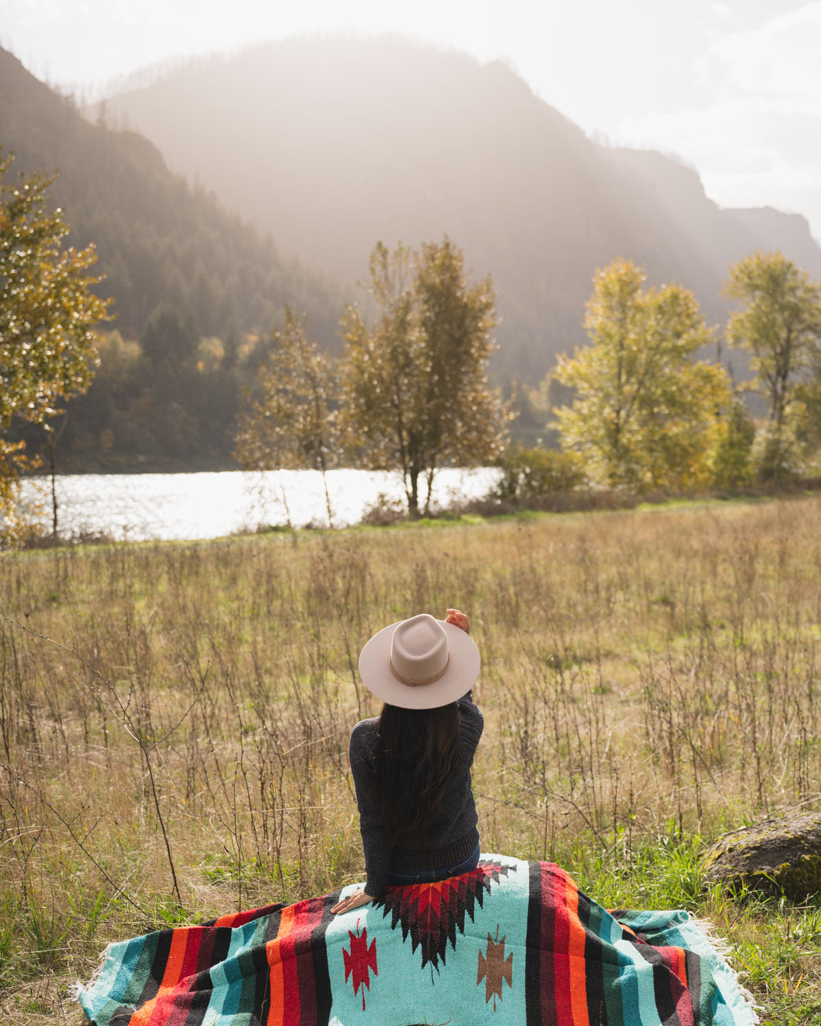 Woman wearing a cowboy hat sitting on a teal and red handwoven mexican diamond blanket in front of sunset in the mountains
