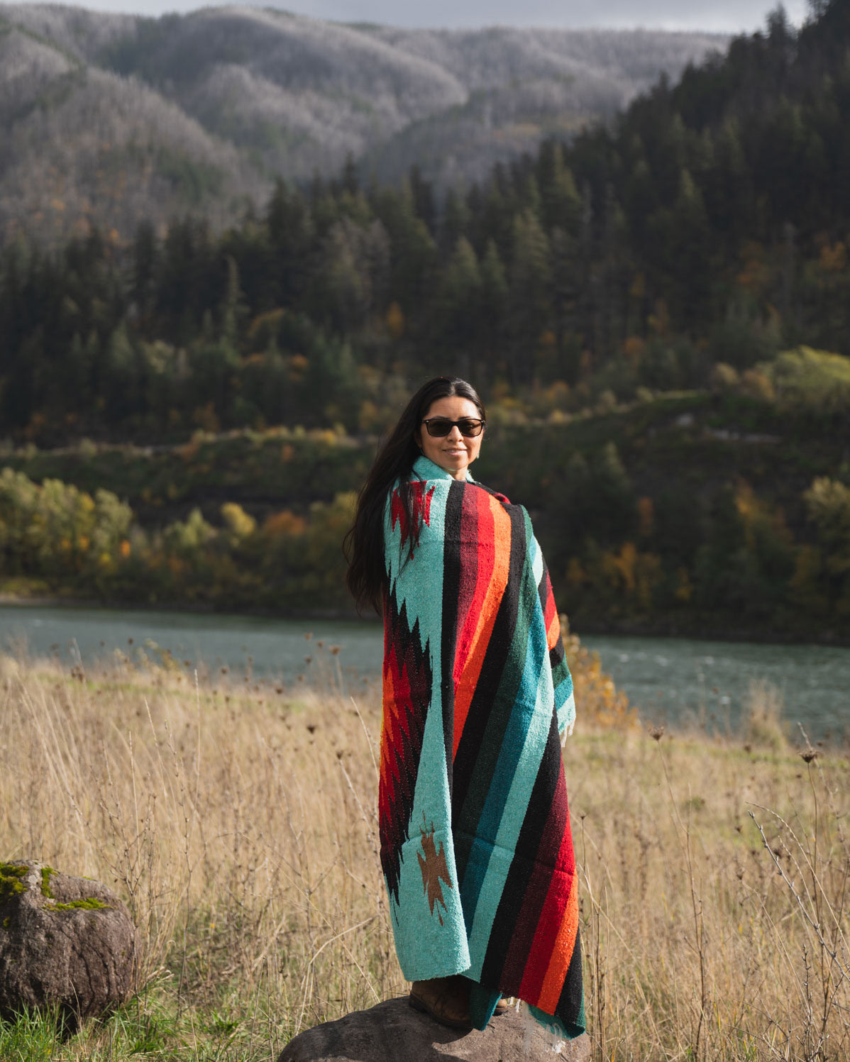 Woman wrapped in a a teal and red handwoven mexican diamond blanket in front of sunset in the mountains