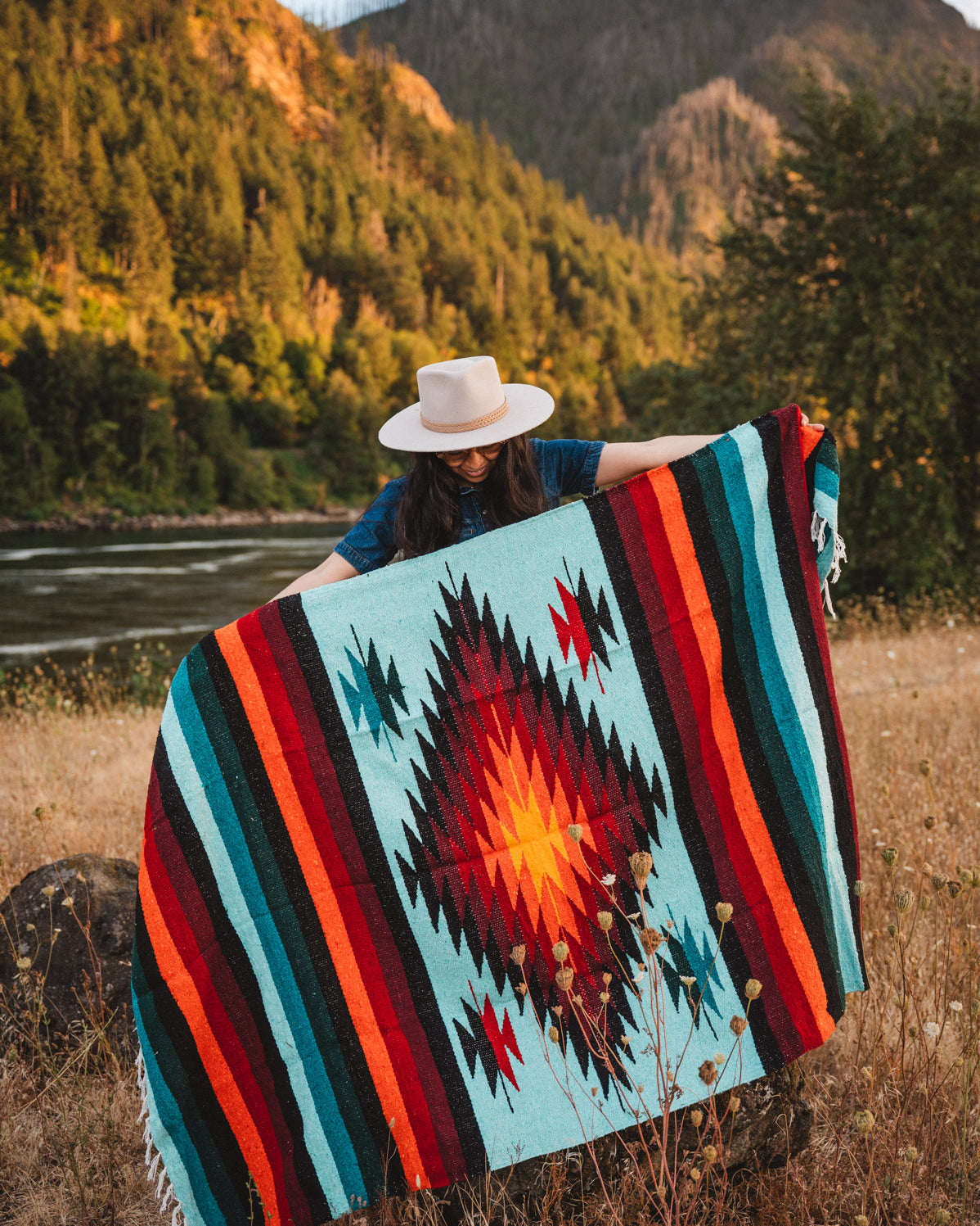 Woman holding teal handwoven diamond mexican blanket in front of mountain at sunset