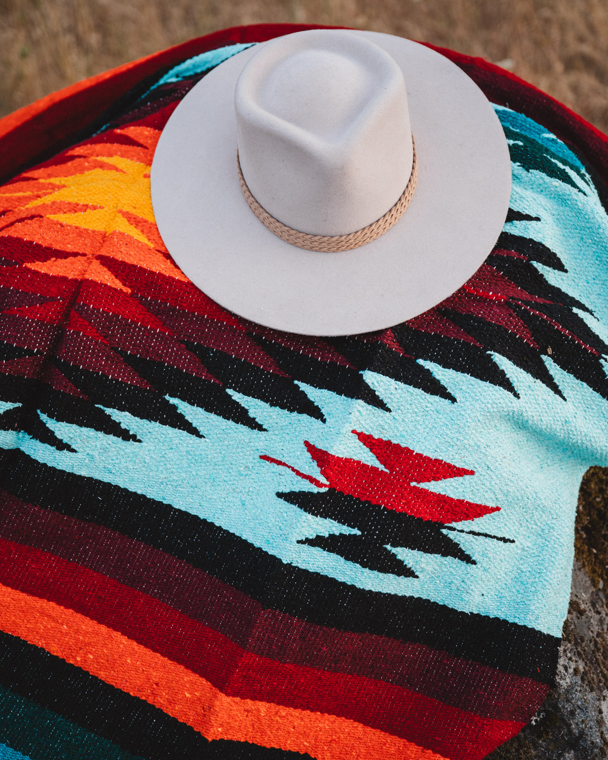 A white cowboy hat with a tan rope band rests on a colorful woven blanket featuring geometric patterns in turquoise, black, red, orange, and dark blue.