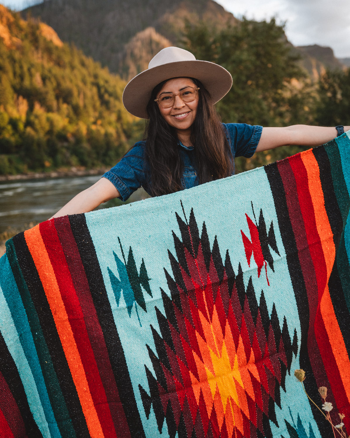 Woman holding teal handwoven diamond mexican blanket in front of mountain at sunset