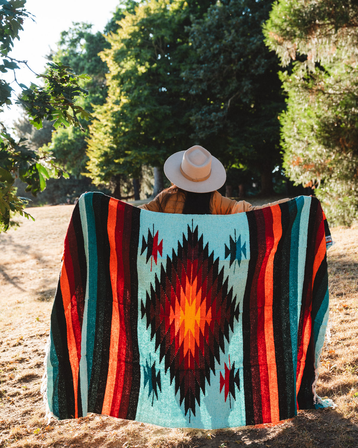 A person sitting outdoors in a wooded area, viewed from behind, wrapped in a large Southwestern-style woven blanket. The blanket features a turquoise background with a large red, orange, and black diamond motif, flanked by vertical stripes of red, orange, black, and teal.