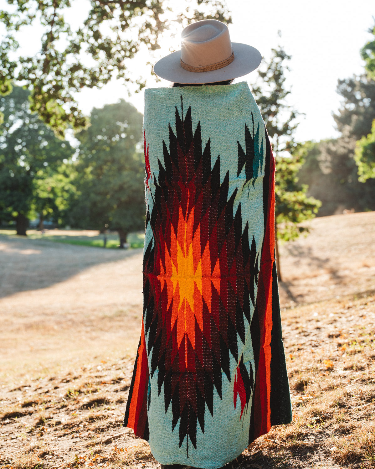 A person standing outdoors in a park wearing a wide-brimmed straw hat and a long woven blanket draped over their body, showing a Southwestern-style geometric pattern in turquoise, black, red, and orange.