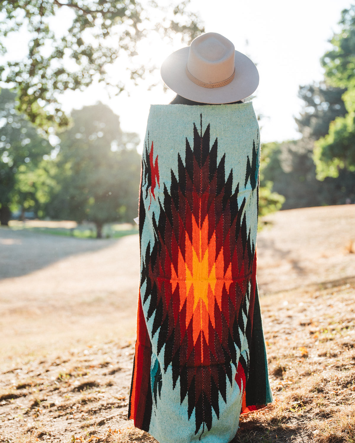 A person standing outdoors in a park, viewed from behind, wearing a wide-brimmed light beige hat and a long woven blanket or wrap with a bold Southwestern geometric pattern in turquoise, black, red, orange, and yellow.