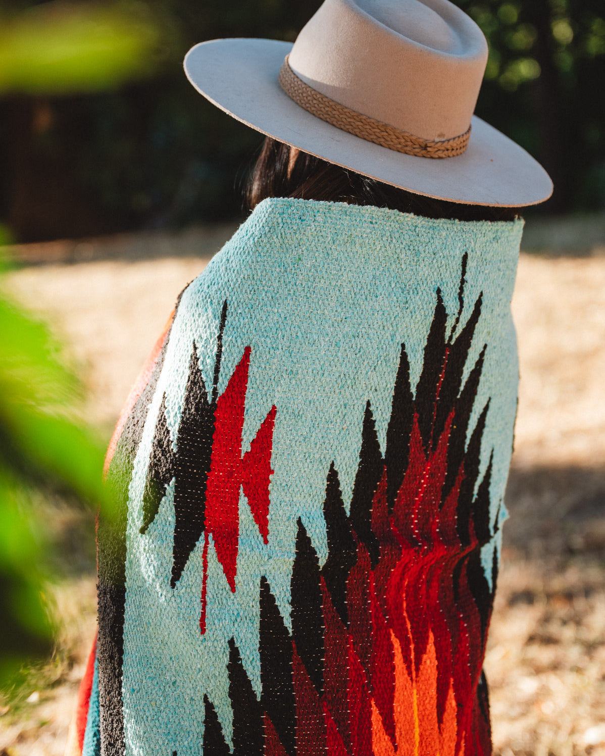 A person outdoors wearing a wide-brim beige hat and a light blue woven blanket or poncho with bold black, red, and orange geometric patterns, shown from the back.
