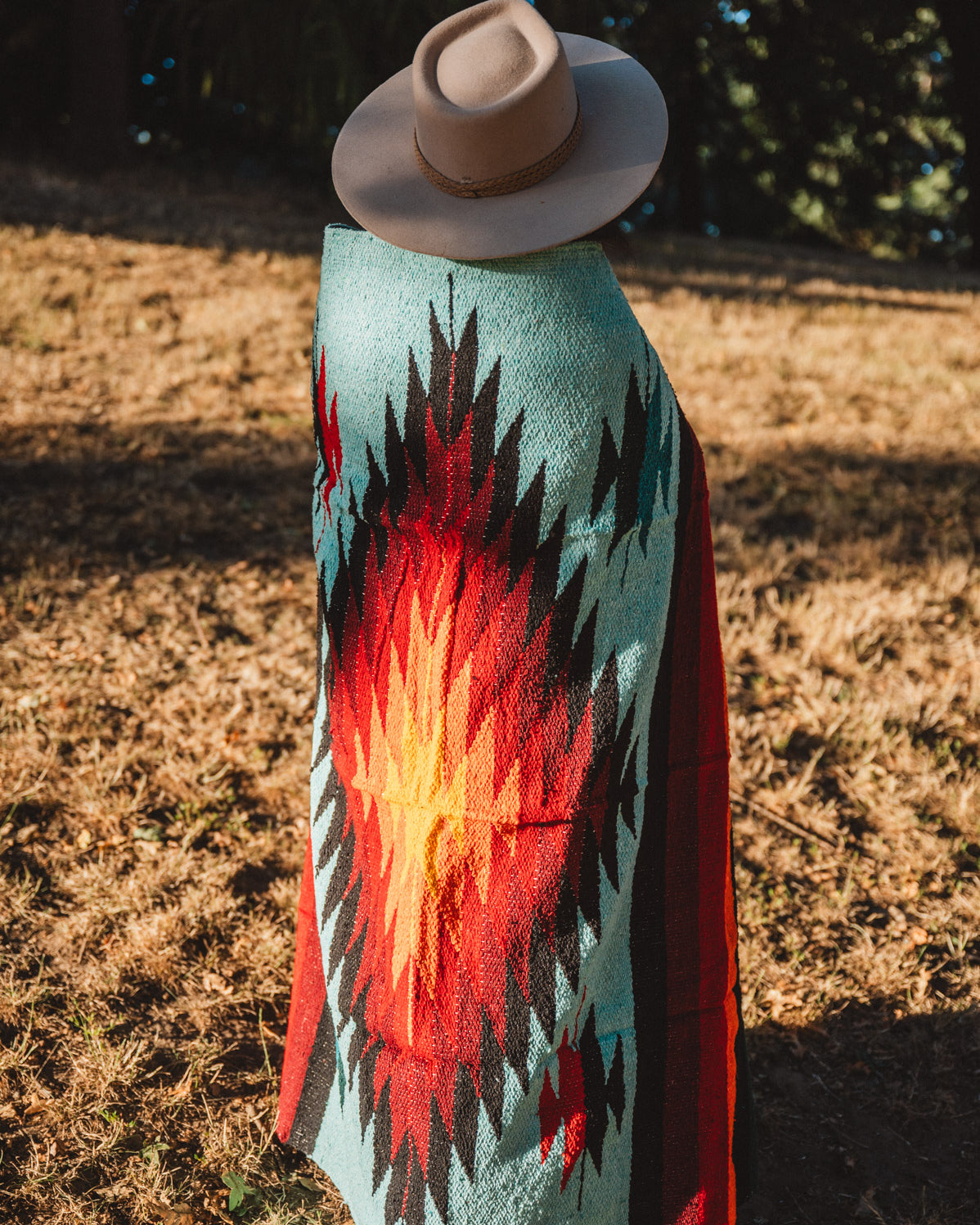 A person standing outdoors in a dry grassy field, viewed from behind, wearing a wide-brim beige hat and a large woven blanket or poncho with a Southwestern-style geometric pattern in turquoise, red, orange, black, and white.