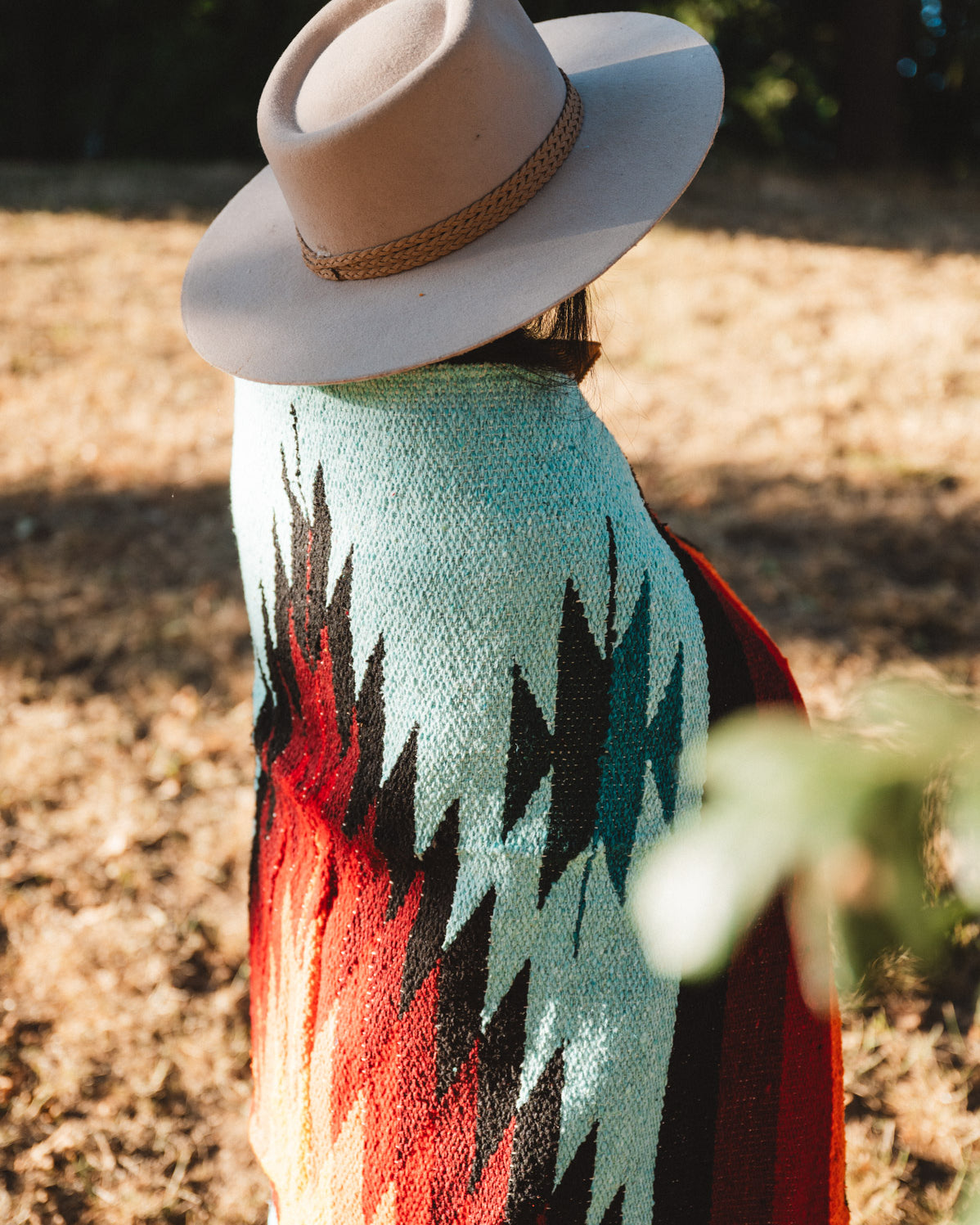 A person outdoors wearing a wide-brim beige hat and a colorful woven blanket or poncho with a Southwestern-style geometric pattern in turquoise, black, red, and orange.