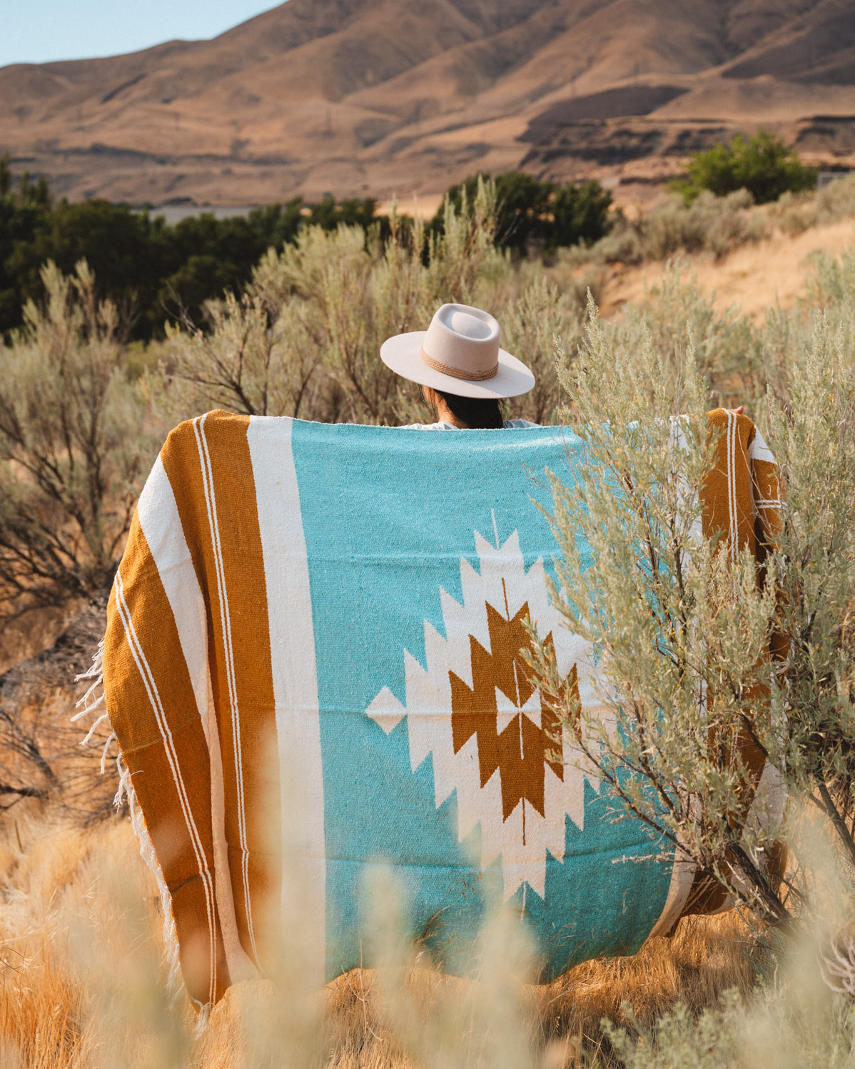 A person wearing a wide-brim hat sits outdoors in a desert landscape, wrapped in a large woven blanket with turquoise, mustard, white, and brown geometric patterns.