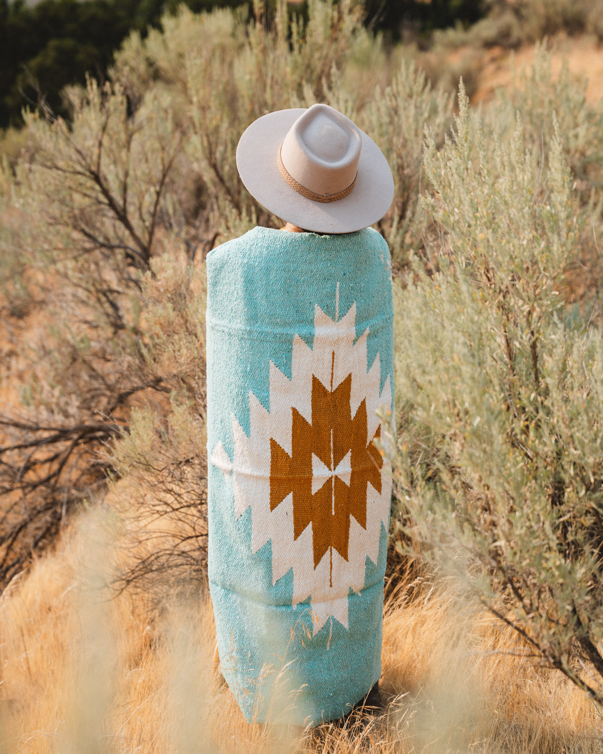 A person standing outdoors in a dry grassy landscape, viewed from behind, wearing a turquoise blanket with a large white and brown geometric Southwestern-style pattern and a light beige wide-brim hat.