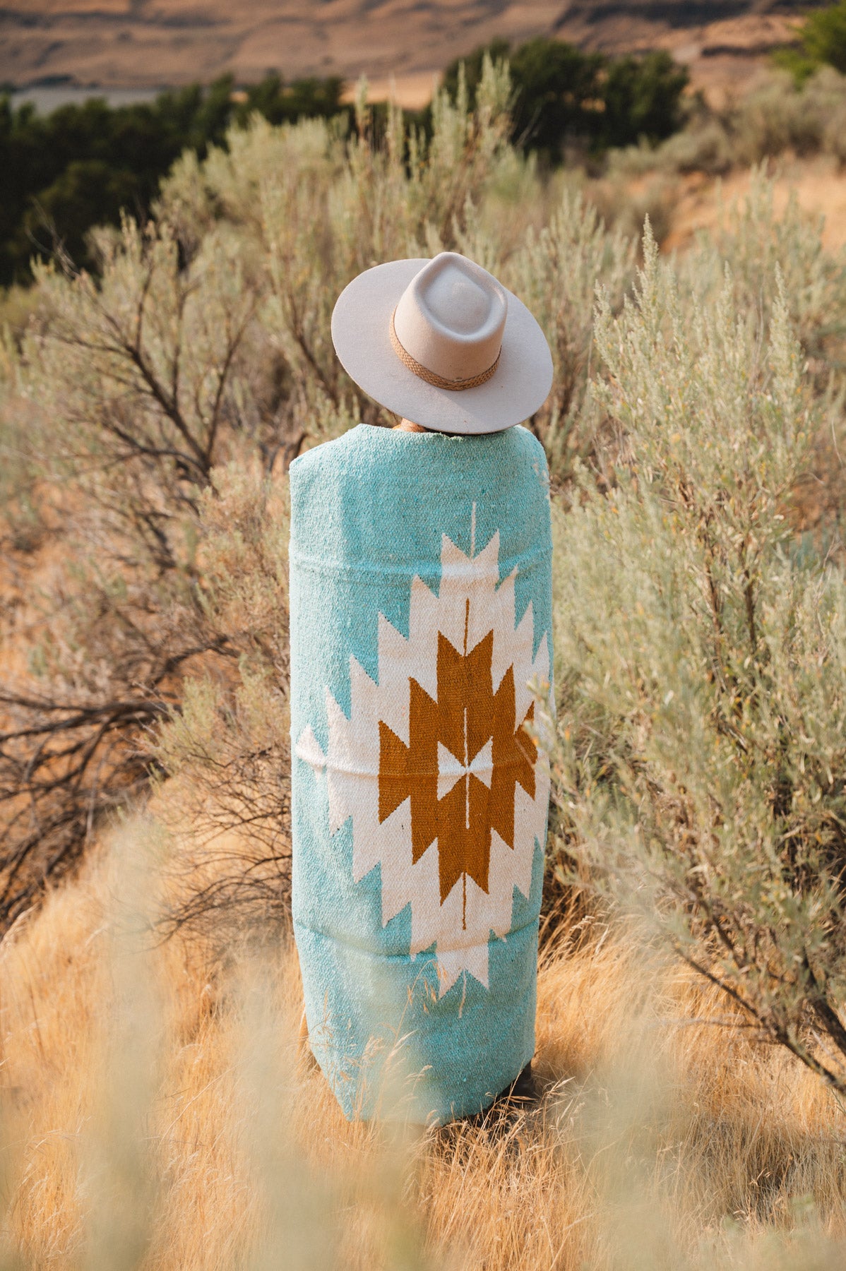 A person standing outdoors in a dry grassy landscape, viewed from behind, wearing a turquoise blanket with a large white and brown geometric Southwestern-style pattern and a light beige wide-brim hat.