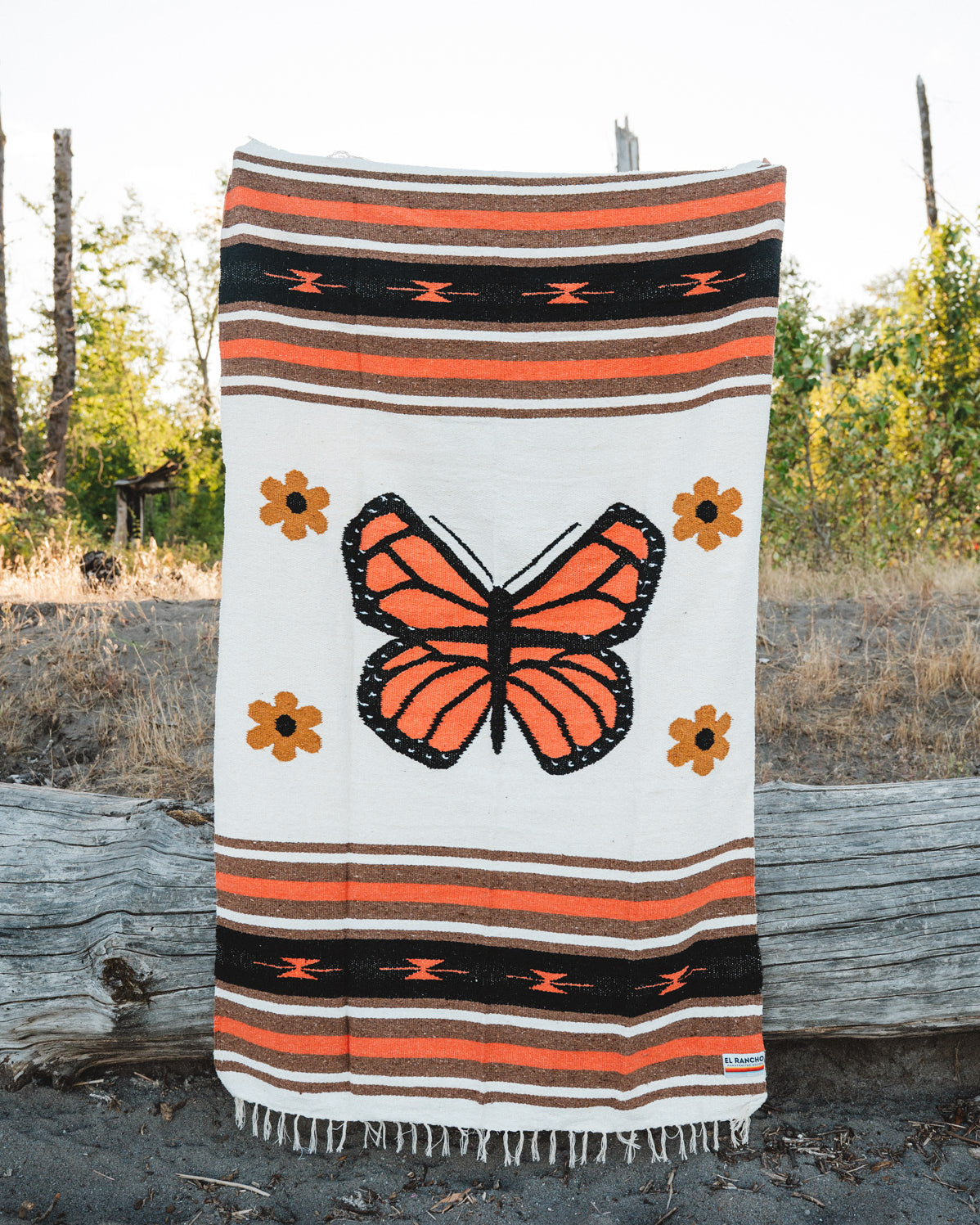 A woven blanket with a large orange and black butterfly design, surrounded by small orange flowers and horizontal striped bands, draped over a log outdoors.