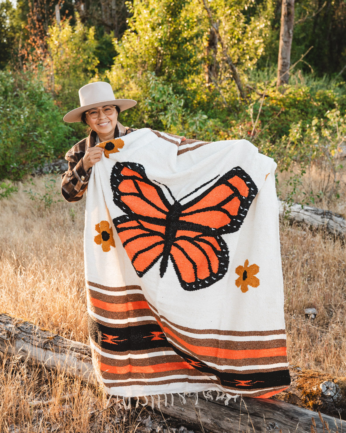 A person outdoors in a wooded, grassy setting holds up a large woven blanket featuring a prominent orange and black butterfly design on a cream background, with orange and brown floral and striped accents.