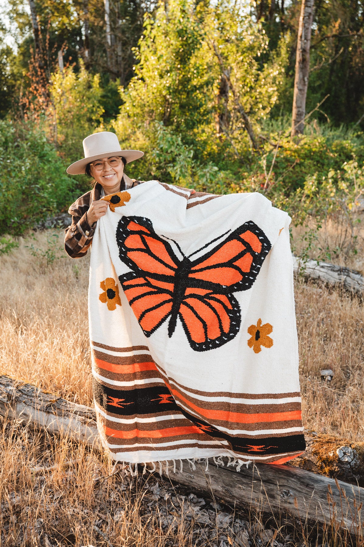 A person outdoors in a wooded, grassy setting holds up a large woven blanket featuring a prominent orange and black butterfly design on a cream background, with orange and brown floral and striped accents.