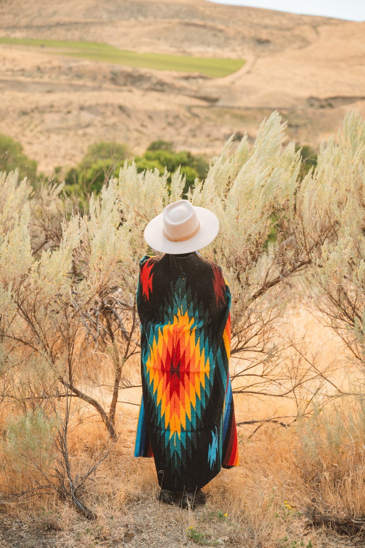 A person viewed from behind wearing a long, vibrant Southwestern-style woven poncho with a red, orange, yellow, and blue geometric diamond pattern, standing in a dry grassy field with shrubs and hills in the background.