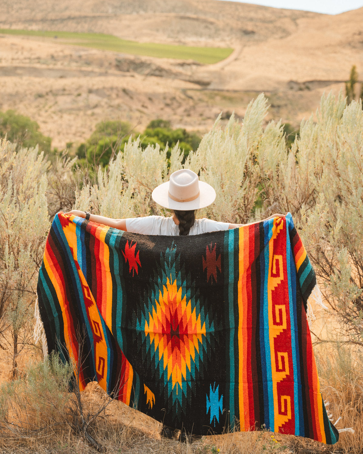 A person standing outdoors in a dry grassy landscape holds up a large woven blanket with a bold Southwestern geometric pattern in black, orange, red, yellow, and blue.