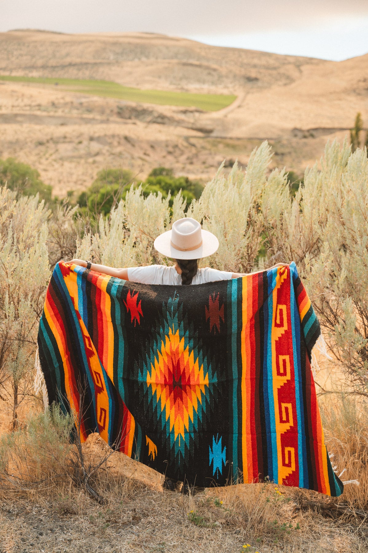 A person standing outdoors in a dry grassy landscape holds up a large woven blanket with a bold Southwestern geometric pattern in black, orange, red, yellow, and blue.