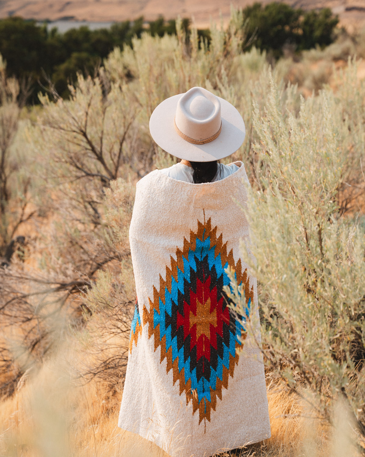 A person stands outdoors in a desert landscape wearing a light beige wide-brim hat and a cream woven poncho with a large Southwestern-style diamond pattern in blue, red, yellow, and brown.
