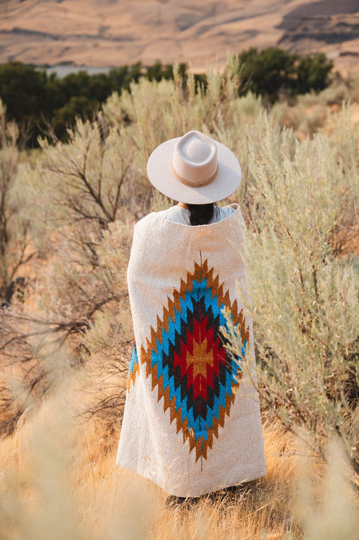 A person stands outdoors in a desert landscape wearing a light beige wide-brim hat and a cream woven poncho with a large Southwestern-style diamond pattern in blue, red, yellow, and brown.