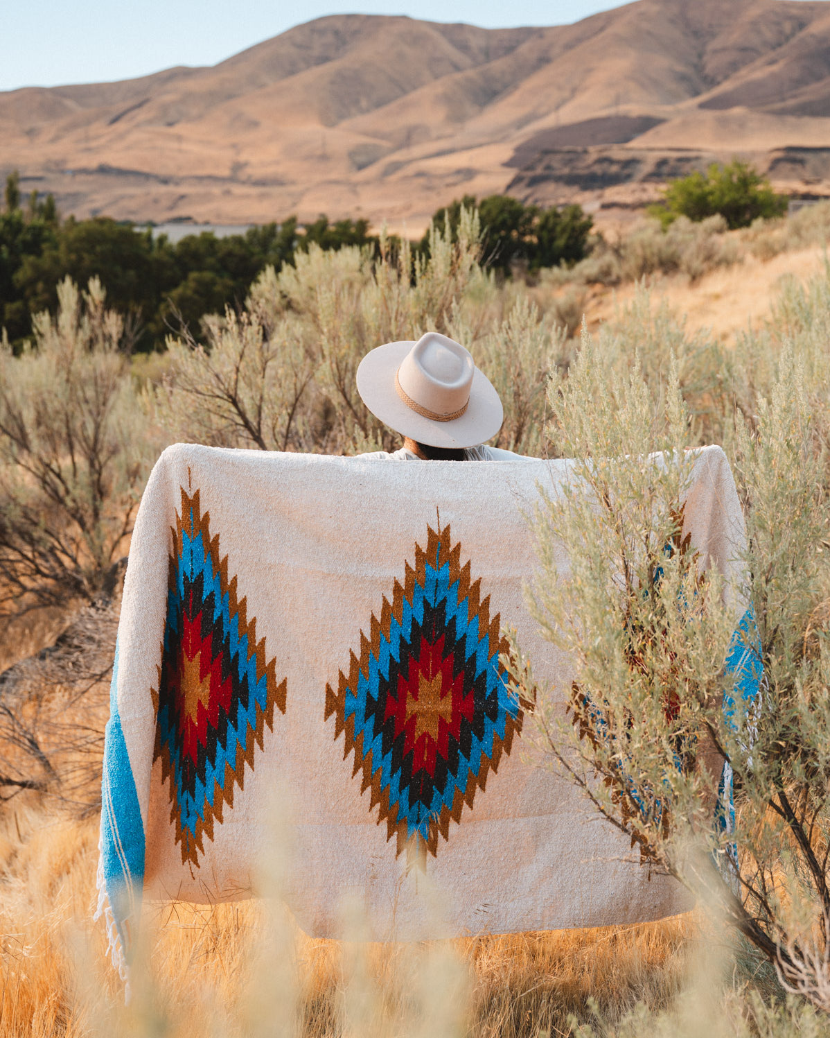 A person seen from behind wearing a wide-brimmed light hat and wrapped in a large southwestern-style blanket with two large diamond motifs in blue, red, brown, and cream, standing in a dry grassy landscape with hills in the background.