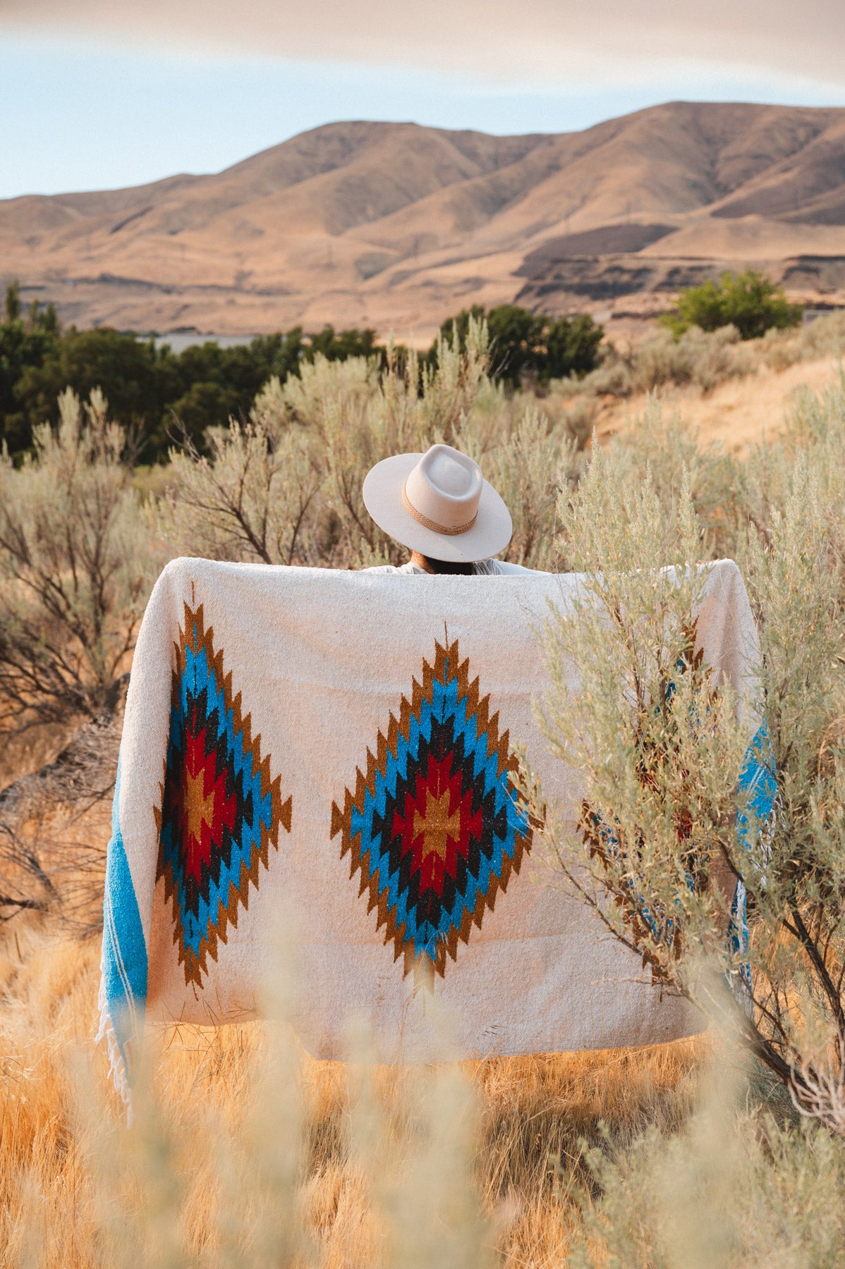 A person seen from behind wearing a wide-brimmed light hat and wrapped in a large southwestern-style blanket with two large diamond motifs in blue, red, brown, and cream, standing in a dry grassy landscape with hills in the background.