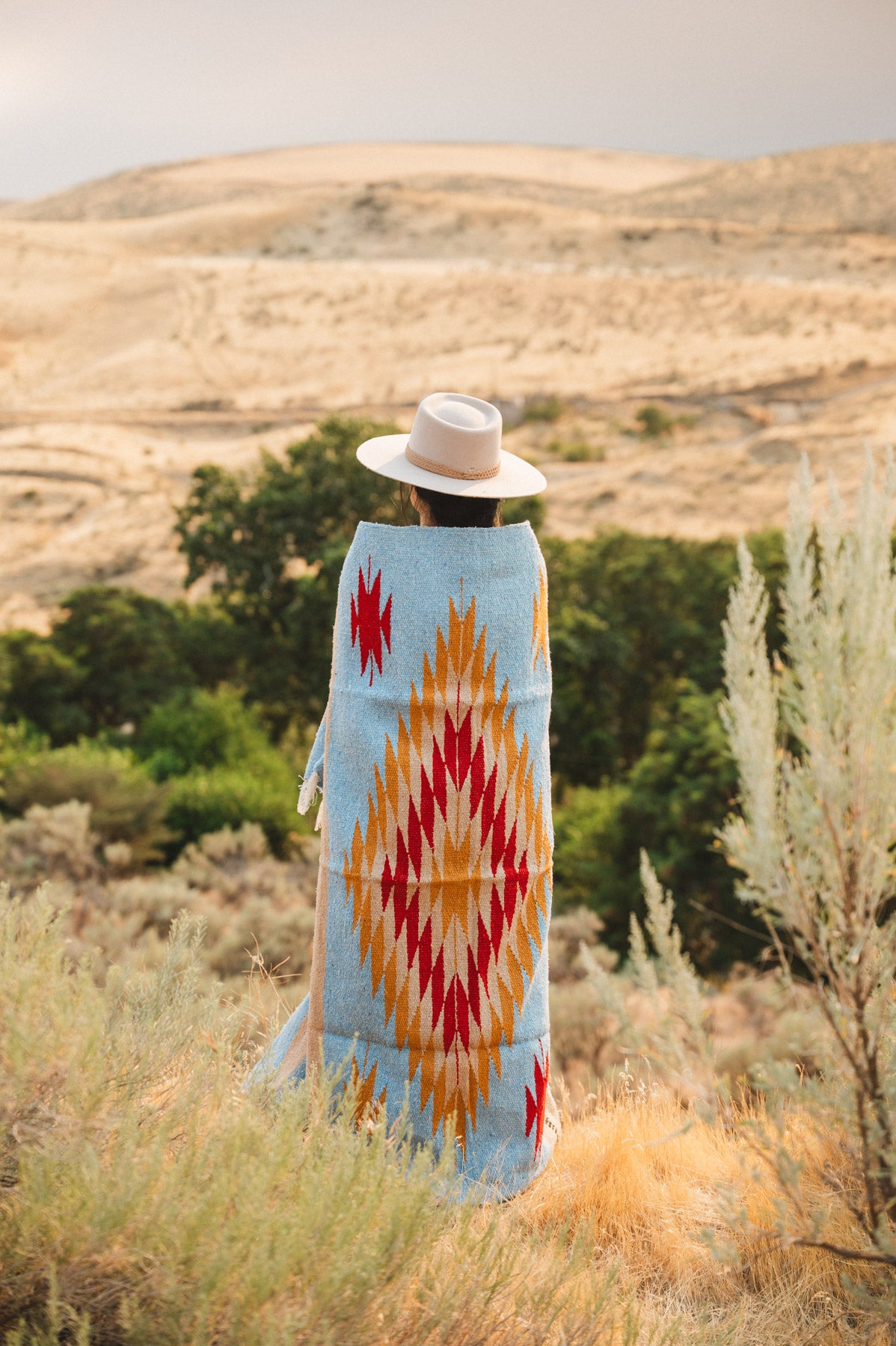 A person standing outdoors in a desert landscape, viewed from behind, wrapped in a light blue blanket with a large red, orange, and yellow geometric diamond pattern and wearing a white hat.