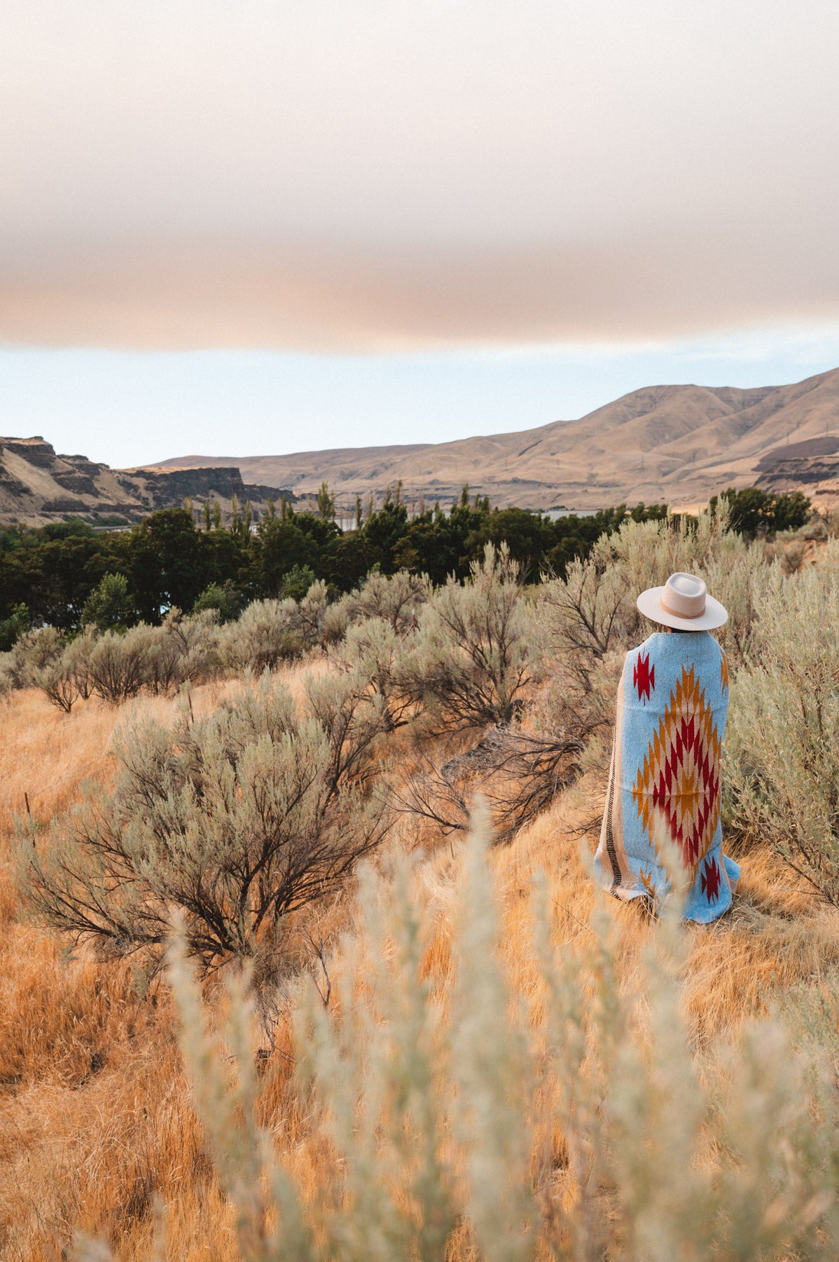 A person stands in a dry grassy landscape wearing a wide-brimmed hat and a light blue blanket or poncho with a large red, yellow, and white geometric diamond pattern, with hills and trees in the background.
