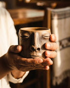 Hand holding a ceramic mug shaped like a face with closed eyes and a neutral expression.