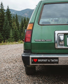 Green Toyota Tercel 4WD with a humorous bumper sticker on a gravel road with trees and mountains in the background.
