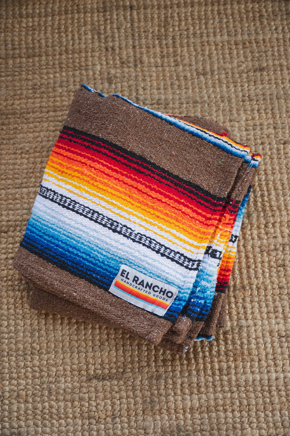 A folded serape blanket with brown borders and a colorful striped pattern of red, orange, yellow, white, and blue, featuring a visible 'EL Rancho' label, displayed on a textured tan woven rug.