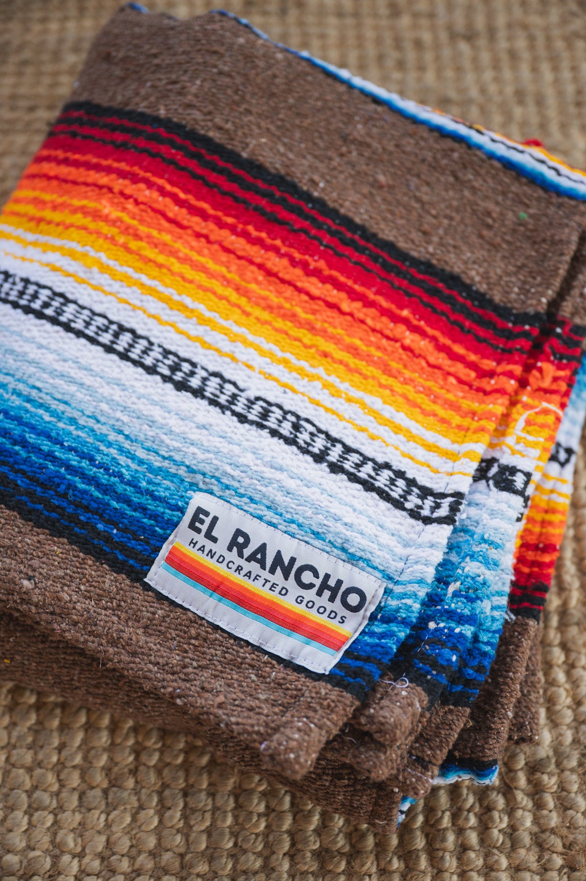 A folded woven blanket with a colorful serape-style striped pattern in brown, black, white, blue, orange, yellow, and red, featuring a visible 'EL RANCHO Handcrafted Goods' label, set on a woven beige surface.