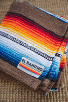 A folded woven blanket with a colorful serape-style striped pattern in brown, black, white, blue, orange, yellow, and red, featuring a visible 'EL RANCHO Handcrafted Goods' label, set on a woven beige surface.