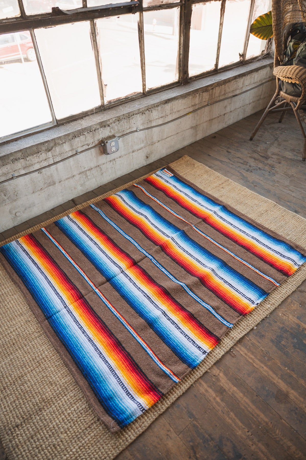 A rectangular woven rug with bold horizontal serape-style stripes in blue, white, orange, red, and brown, placed on a natural jute mat over a dark wood floor in a loft-style room with large windows.