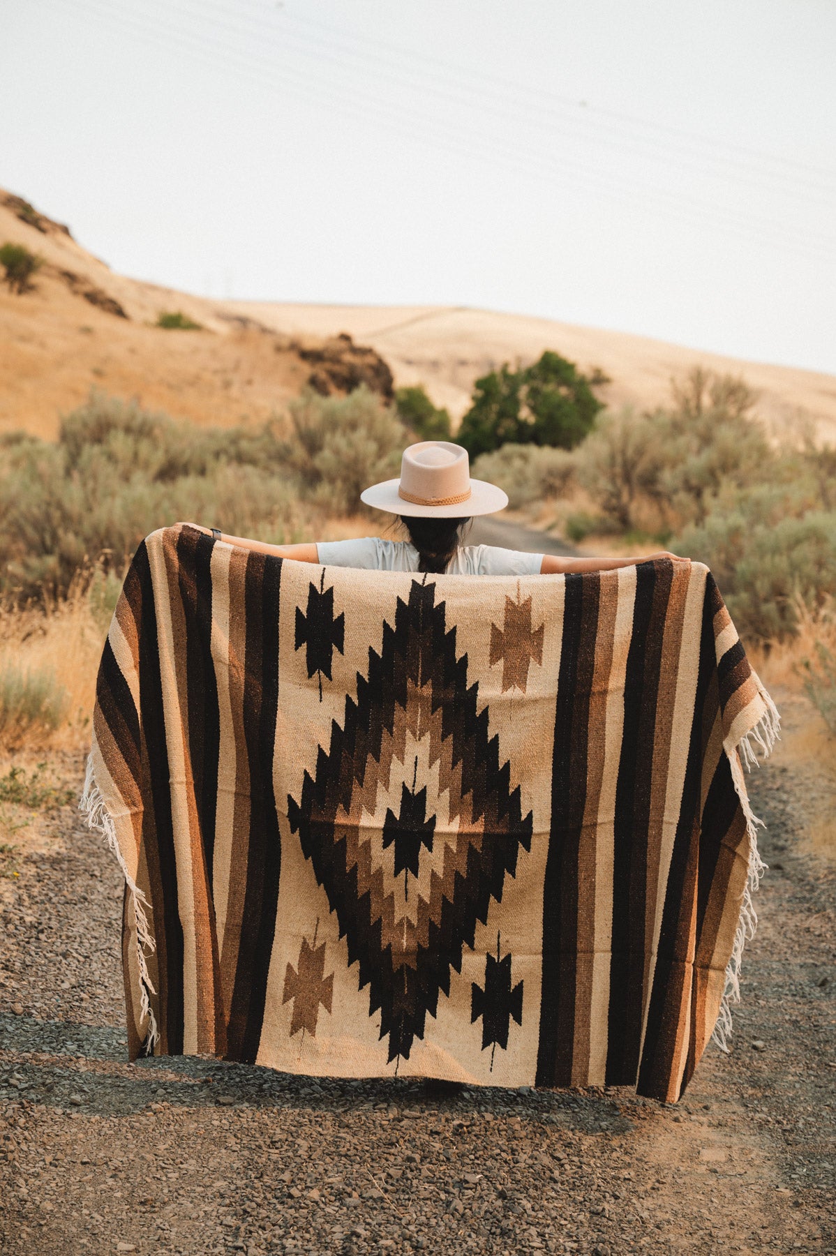 A person standing outdoors on a dirt path in a desert landscape, holding up a large woven blanket or poncho with a Southwestern geometric pattern in beige, brown, and black. The person is wearing a wide-brimmed hat.