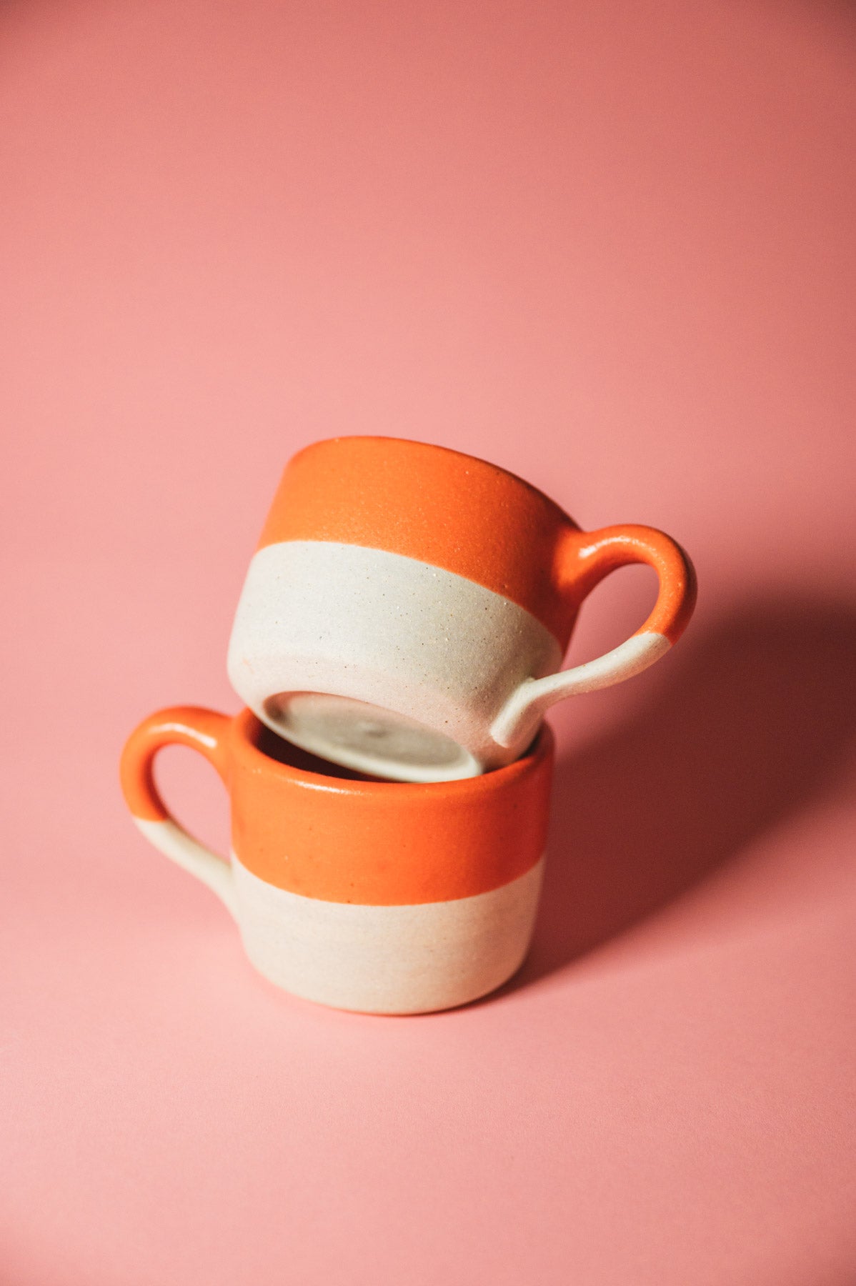Two orange and white ceramic cups stacked on a pink background