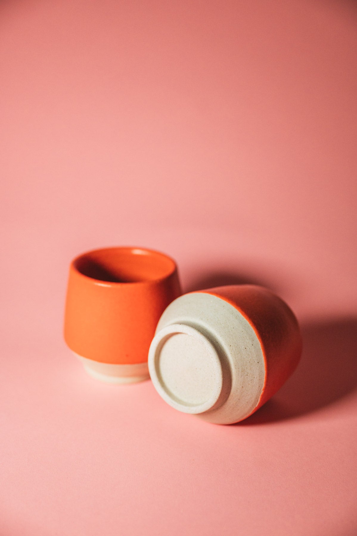 Two ceramic cups, one orange and one beige with white interior, on a pink background