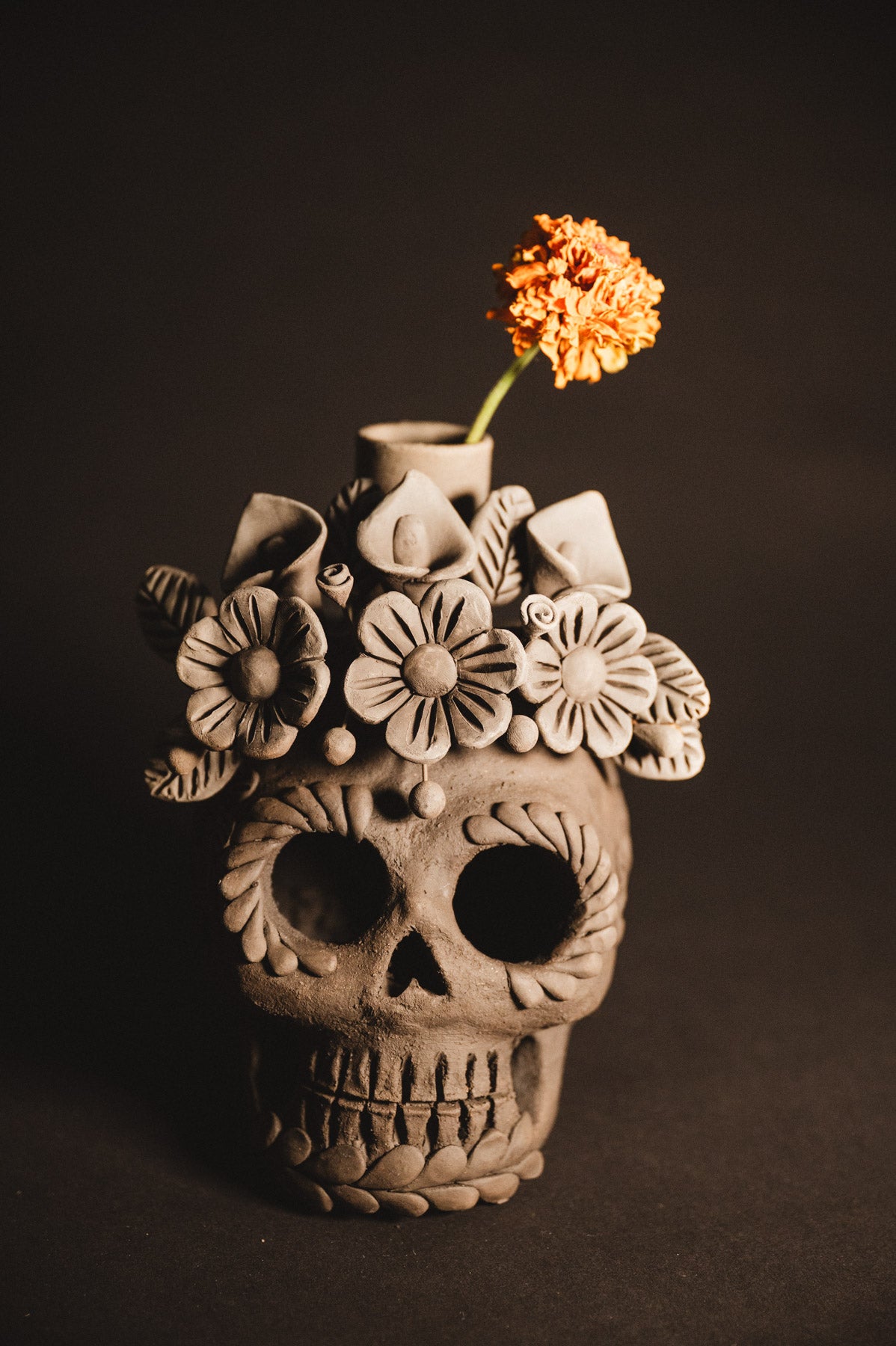 Ceramic skull vase with floral designs and a single orange flower on a dark background