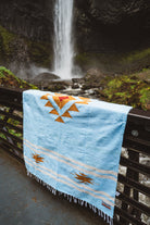 Blue quilt with orange and yellow patterns draped over a railing in front of a waterfall.