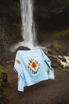 Light blue blanket with a geometric pattern draped over a rock in front of a waterfall.