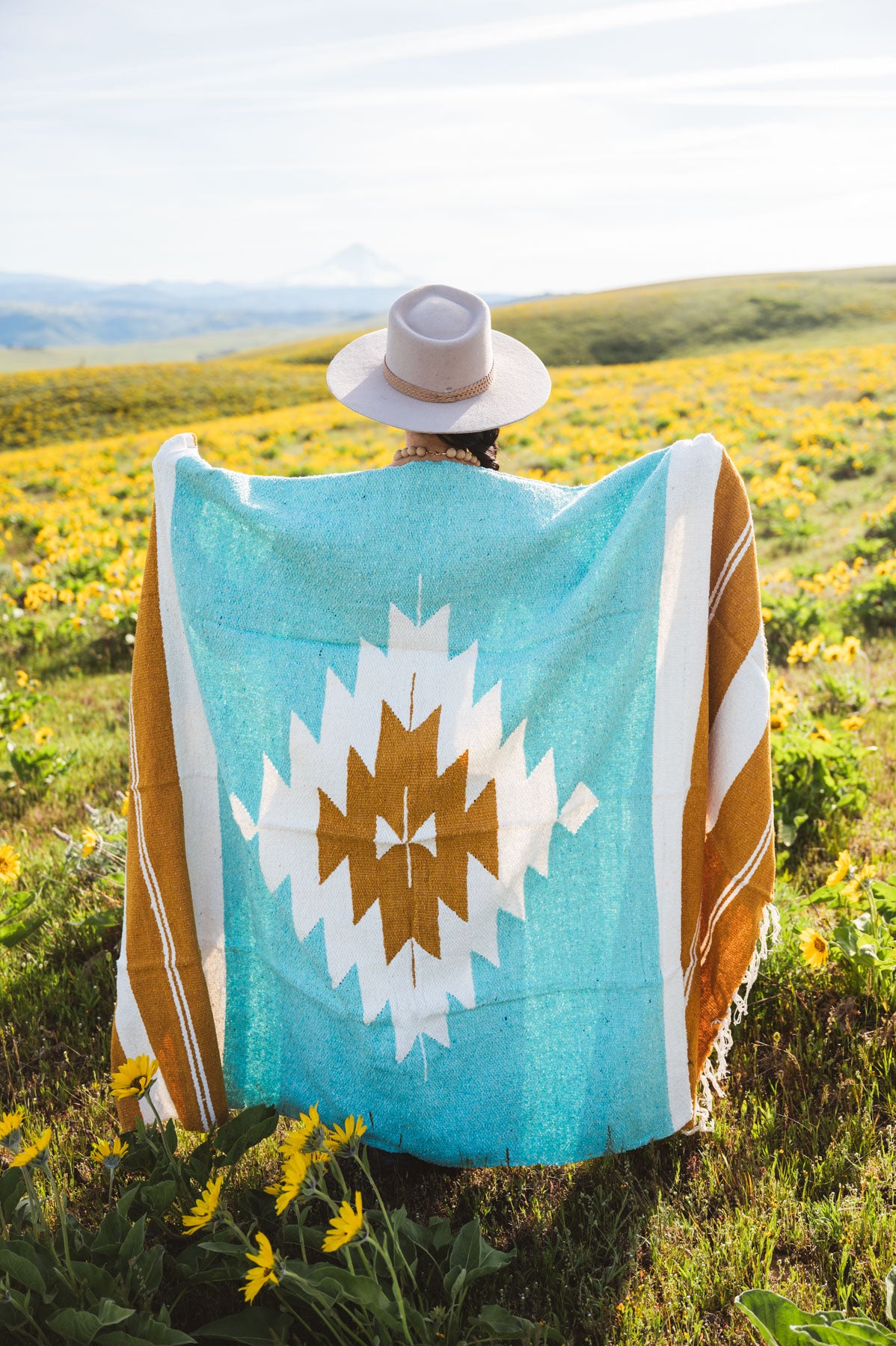 Person wearing a turquoise blanket with a white and brown pattern in a field of yellow flowers.