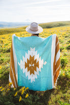 Person wearing a turquoise blanket with a white and brown pattern in a field of yellow flowers.