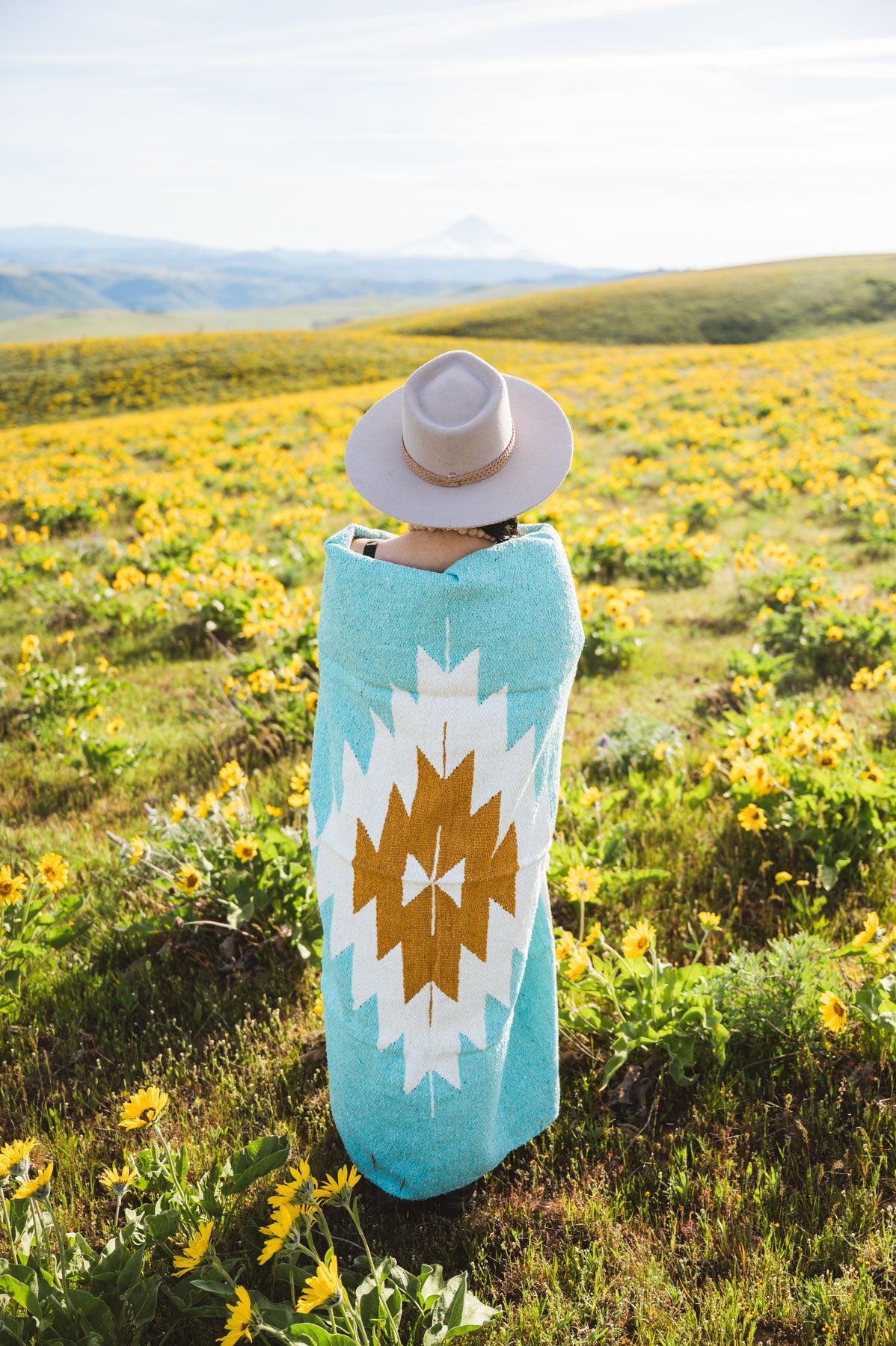 Person wrapped in a turquoise towel with a white and brown pattern, standing in a field of yellow flowers.