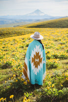 Person wearing a turquoise blanket with a white and brown diamond pattern in a field of yellow flowers with mountains in the background.