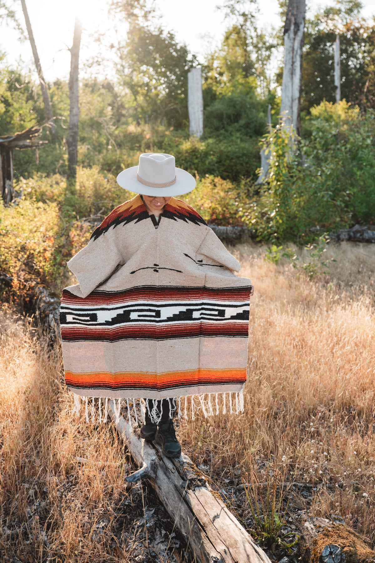 A person standing outdoors in a dry grassy field wearing a wide-brimmed white hat and a large woven poncho with geometric black, red, orange, and beige patterns and fringe.