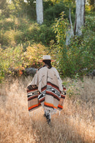 A person viewed from behind walking through tall dry grass in a wooded outdoor setting, wearing a wide-brim hat and a beige woven poncho with black, white, and orange geometric pattern details.