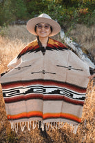 A person outdoors wearing a wide-brim hat and a large woven poncho with geometric Southwestern-style stripes in beige, black, red, orange, and white, standing in dry grass.