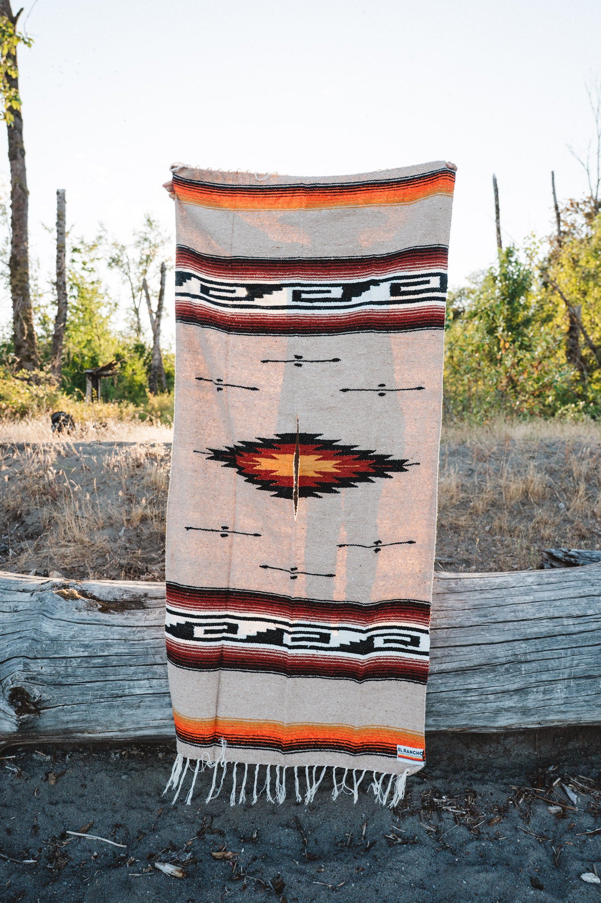 A Southwestern-style woven blanket with fringe is draped over a weathered log outdoors, with trees and dry grass blurred in the background.