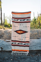 A Southwestern-style woven blanket with fringe is draped over a weathered log outdoors, with trees and dry grass blurred in the background.