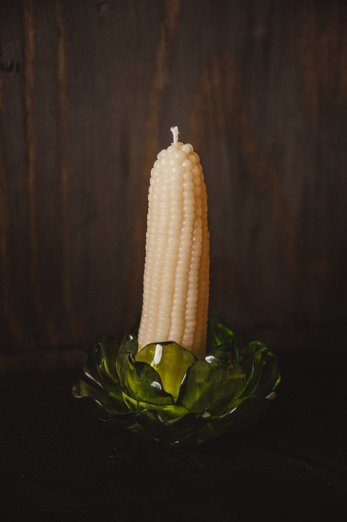 An off-white corn cob pillar candle standing upright in a translucent green glass holder against a dark wooden background.